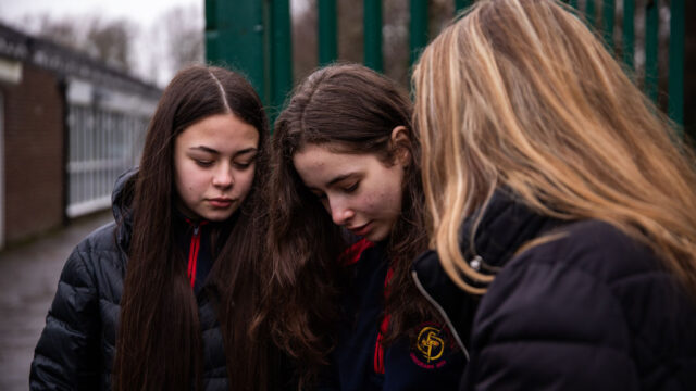 This is a photo of three girls outside of a school, all looking at a mobile phone.