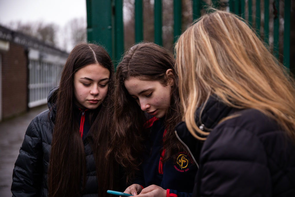 This is a photo of three girls outside of a school, all looking at a mobile phone.